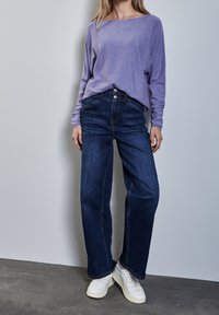 Lila långärmad tröja, löst passande, med vid halsringning. Mörkblå högmidjade jeans, rak ben, med klassisk midjoknapp. Vita sneakers.