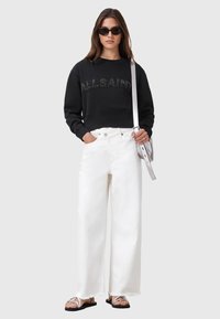 Felpa nera corta con testi "ALL SAINTS", jeans bianchi a gamba larga e sandali neri. Una borsa argentata e occhiali da sole completano il look.