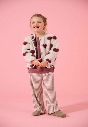 Ung pige smilende, iført en hvid fleecejakke med mørke hjerter, mørkerød bluse, stribede bukser og brune sko, stående på en lyserød baggrund.