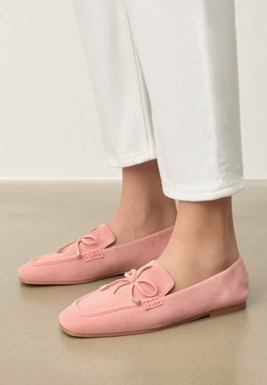 Füße, die hellrosa Wildleder-Loafer mit dekorativer Schleife und Ausschnittdetail tragen, kombiniert mit weißen, gekürzten Hosen auf einem glatten beigen Boden.