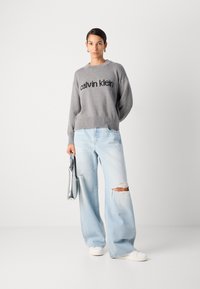 Suéter gris con el logo "calvin klein", jeans de pierna ancha de color azul claro con detalle desgastado, zapatillas blancas y un bolso de charol plateado.