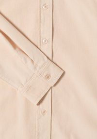 Camicia button-up di un leggero beige, realizzata in tessuto a coste. Presenta un colletto ampio, maniche lunghe con polsini e cinque bottoni abbinati lungo la chiusura.