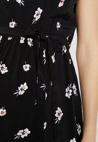 Robe noire avec petits motifs floraux blancs et roses, dotée d'un ruban noué à la taille et d'un tissu froncé sous la taille, portée sur un bras nu.