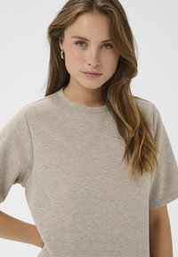 Beidzša īsām piedurknēm svīteris bēšā krāsā ar apaļu kaklu, mīksta tekstūra un brīva piegriezuma, attēlots uz vienkārša balta fona.