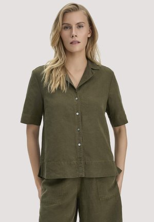 Blonde Frau mit olivgrünem, kurzärmeligem Hemd mit Knöpfen und passender kurzer Hose, steht mit den Händen in den Hosentaschen vor schlichtem Hintergrund.