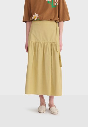 Jupe portefeuille beige avec détail froncé, associée à un top à manches courtes marron à motif floral et des chaussures plates tissées crème portées par une personne.