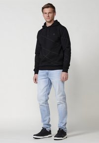 Hanorac negru cu glugă, cu un model diagonal, purtat cu blugi de un albastru deschis și adidași negri, având un design simplu și un material texturat.