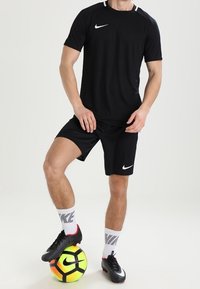 Musta urheilupaita ja -shortsit, joissa on valkoinen Nike-logo, yhdistetty mustiin jalkapallokenkiin ja valkoisiin sukkiin. Pelaaja seisoo värikkään jalkapallon pä�ällä.