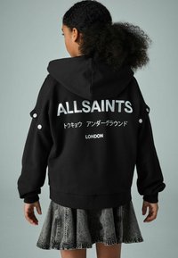 Crna oversized hoodie sa sljedećim natpisima izrađenim od rhinestona i grafičkim printovima na stražnjoj strani, uparen s teksturiranom traper suknjom na širenje.