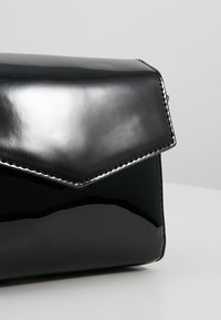 Borsa a pochette in pelle verniciata nera con chiusura a patta geometrica, accentuata da cuciture argentate lungo il bordo, finitura lucida e design elegante.