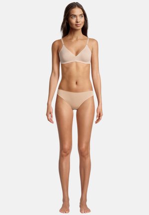 Donna in piedi a piedi nudi che indossa un reggiseno senza cuciture beige e slip abbinati su sfondo bianco semplice.