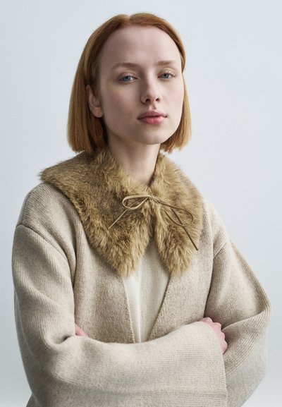 Μπεζ ζακέτα με φαρδιά γιακά από μαλακή, ψεύτικη γούνα σε ανοιχτό καφέ τόνο και δεμένη με λεπτή καφέ κορδέλα. Απλή πλεκτή υφή.