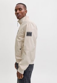 Veste beige avec un col montant, fermeture éclair, poches latérales, et un patch logo noir sur la manche. Le matériau semble lisse et léger.