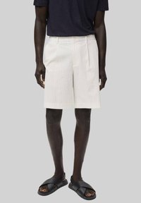 Weiß gestreifte Shorts mit geradem Schnitt, ausgestattet mit einem Reißverschluss vorne und zwei seitlichen Taschen, getragen mit schwarzen Rutsch-Sandalen.