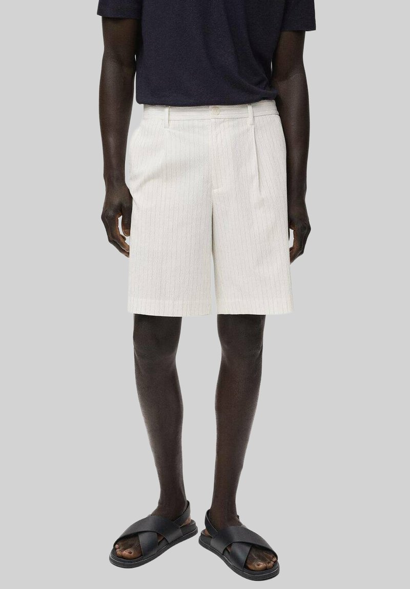 Weiß gestreifte Shorts mit geradem Schnitt, ausgestattet mit einem Reißverschluss vorne und zwei seitlichen Taschen, getragen mit schwarzen Rutsch-Sandalen.