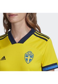 Gul fodboldtrøje med en marinefarvet krave og striber, som har et blåt og gult svensk våbenskjold på brystet og Adidas-logo på siden.