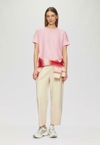 Rosa gestreiftes T-Shirt mit kurzen Ärmeln, kombiniert mit beigen, weit geschnittenen Hosen. Ein gestrickter, mehrfarbiger Wickel ist um die Taille gebunden. Legere Sportschuhe.