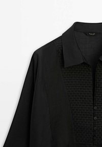 Chemise noire à manches longues avec un panneau à motif carré texturé, boutonnée à l'avant, avec un col classique, présentée sur fond blanc.