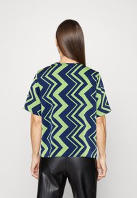 Donna con capelli lunghi e castani che indossa una maglietta a maniche corte con un motivo a zigzag verde e blu navy, abbinata a pantaloni in pelle nera, vista da dietro.
