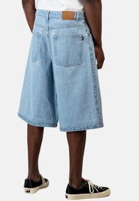 Pantalones cortos de mezclilla azul claro con un corte relajado, que presentan una etiqueta en la cinturilla y dos bolsillos traseros, combinados con zapatillas negras.