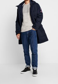 Granatowa parka z kapturem, szary teksturowany sweter, niebieskie dżinsy oraz czarne sneakersy z białymi podeszwami. Na rękawie znajduje się logo.