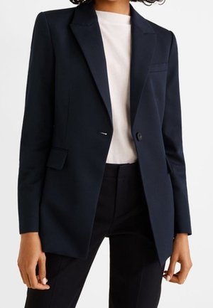 Frau trägt ein maßgeschneidertes dunkelblaues Sakko über einem weißen Top und schwarzen Hosen, steht vor einem schlichten weißen Hintergrund.