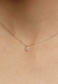 Collar de cadena de plata con un pequeño colgante en forma de lágrima con un detalle brillante, descansando sobre un cuello desnudo. Tono de piel visible en el fondo.