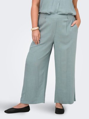Femme portant un pantalon large ample bleu clair avec des poches, des ballerines noires tissées, et un bracelet beige épais, debout avec une main dans la poche.
