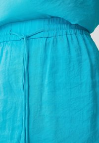 Vêtement turquoise avec une taille élastique froncée, fermeture à cordon, et un tissu doux et texturé. Dispose de poches latérales pour une utilisation pratique.