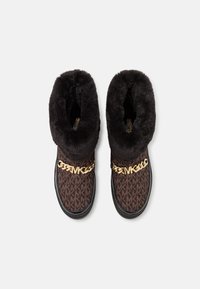 Bruine slippers met een patroon en een voering van nepbont, met een gouden kettingaccent aan de voorkant en een zwarte rubberen zool.