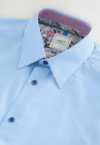Camicia da uomo azzurra chiara con motivo a fantasia, tessuto strutturato, collo con bottoni e foderatura interna del colletto con stampa floreale. Etichetta visibile.