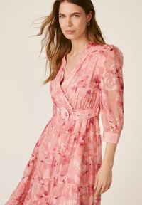 Mujer con cabello largo que lleva un vestido cruzado rosa con flores, mangas abullonadas y un cinturón a juego con hebilla redonda.