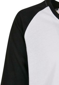 Kortærmet raglan skjorte med sorte ærmer og hvidt kropsstof, lavet af bomuld, der fremhæver en glat tekstur og kontrastsyninger.
