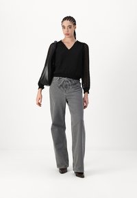 Blouse noire avec un motif de points texturé et des manches légèrement bouffantes, associée à un jean gris à jambes larges et un sac à épaule noir.