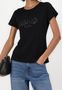 Czarna koszulka z krótkim rękawem z błyszczącym logiem z kryształków "LIU JO MILANO" na środku. W połączeniu z niebieskimi dżinsami i czarną torbą.