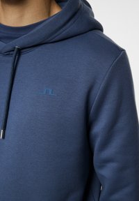 Blå huvtröja gjord av mjukt material, med dragsko i huvan och en liten, matchande logotyp på bröstet. Slät textur, åtsittande design.