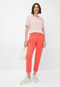 Koraalkleurige katoenen broek met een rechte pasvorm, gecombineerd met een wit en rood gestreept overhemd met korte mouwen en witte sneakers. Houdt een kleine witte tas vast.