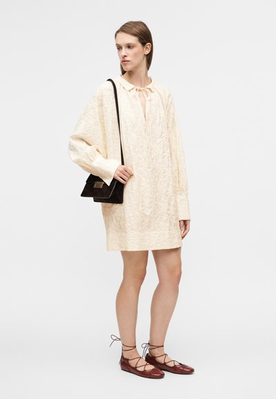 Φόρεμα σε ανοιχτό κρεμ χρώμα με γιακά, υφασμένο με υφή και δέσιμο στο λαιμό, συνδυασμένο με μπορντό δερμάτινα παπούτσια με κορδόνια και μικρή μαύρη τσάντα χιαστί.