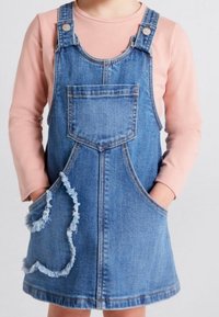 Denim overall dress, vaaleansininen, jossa on iso etutasku, sivutaskut ja sydämen muotoinen säpäleinen taksihyvä. Pitkähihaisessa vaaleanpunaisessa paidassa alla.