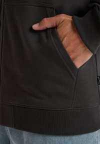 Mørkegrå hoodie med lynlås, der har ribbede manchetter og kant, hånd i lommen, glat tekstur og synlige syninger.