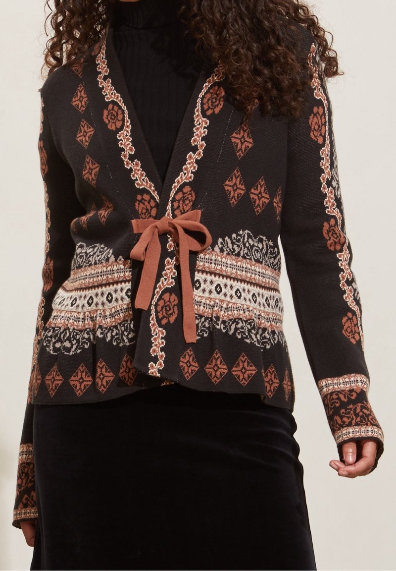 Femme portant un cardigan noir et marron à motif floral, noué avec un ruban marron par-dessus un haut noir et une jupe noire.