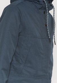 Veste bleu foncé résistante à l'eau avec capuche, cordons contrastants, boutons-pression et détails cousus.