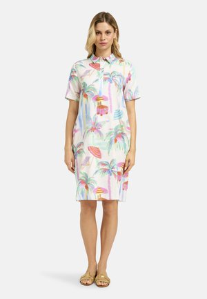 Frau trägt ein knielanges Kurzarmkleid mit farbenfrohem Palmen- und Strandschirm-Print, kombiniert mit beigen Sandalen mit offenen Zehen.