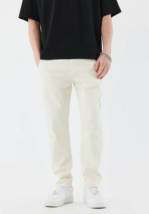 Pantalons beige clair avec une coupe droite, fabriqués en coton, associés à des baskets blanches. Présente un design simple et une taille classique.