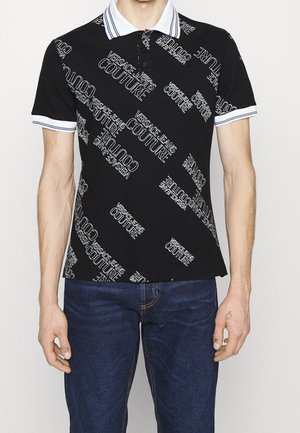 Mand iført sort poloshirt med hvide "VERSACE JEANS COUTURE" tekstmønstre og krave og ærmer med hvide kanter, kombineret med blå jeans.