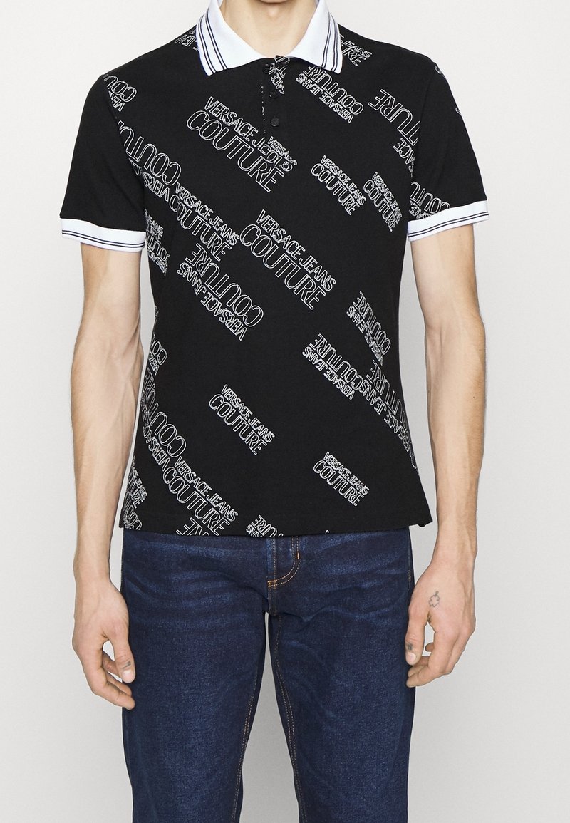 Homme portant un polo noir avec des motifs de texte blancs "VERSACE JEANS COUTURE" et un col et des manches bordés de blanc, associé à un jean bleu.