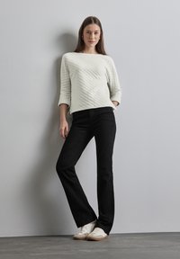 Weißer, gesteppten Pullover mit 3/4-Ärmeln, kombiniert mit schwarzen, ausgestellten Jeans und weißen Sneakers mit beigen Sohlen, vor einem schlichten Hintergrund.