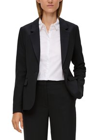 Schwarzer Blazer mitReverskragen, zwei Knöpfen und Seitentaschen. Getragen über einem weißen Hemd mit Kragen, das eine taillierte Passform und glatte Textur aufweist.