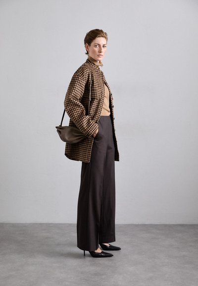 Braun karierter Übergröße-Jacke, beige Oberteil, dunkelbraune weite Hosen und schwarze spitze Absätze. Braune Umhängetasche.