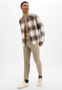 Chemise à carreaux marron et crème, col roulé beige, et pantalon beige slim. Baskets blanches avec des accents marron clair.